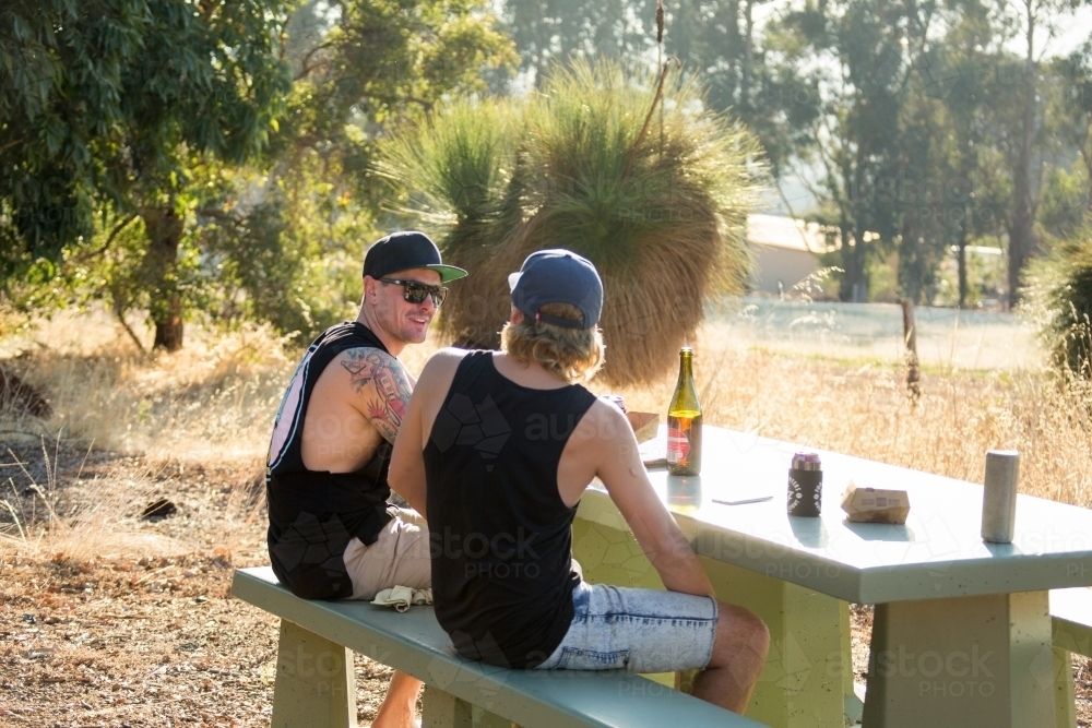 Two young blokes sitting drinking at picnic table outside - Australian Stock Image
