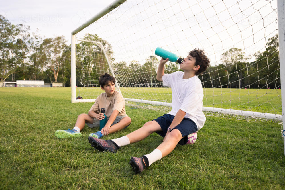 Two young boys drink from their water bottles while sitting on grassy field in front of soccer goal - Australian Stock Image