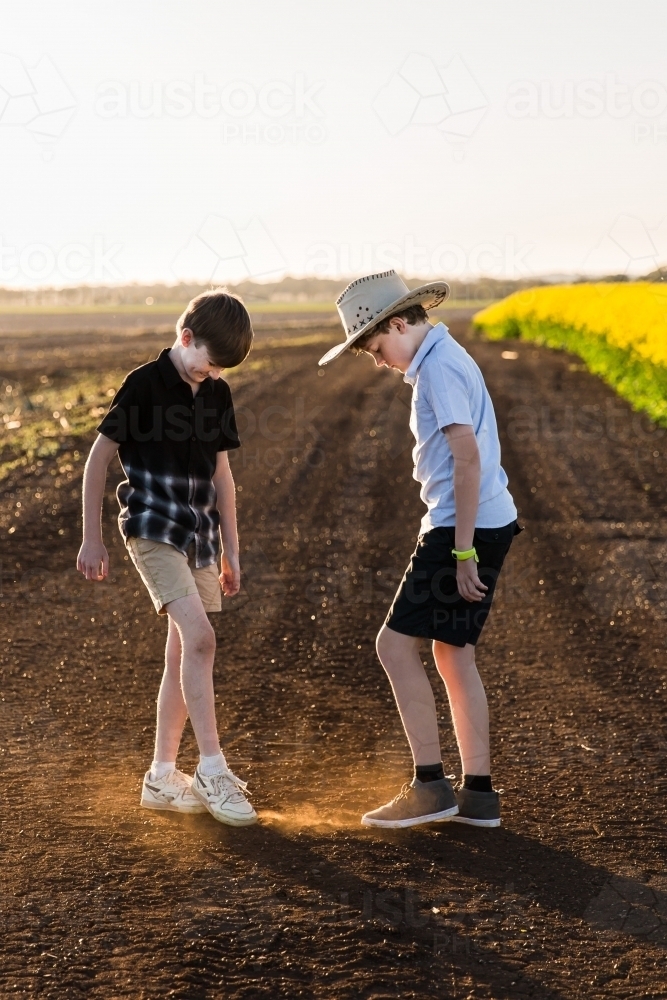 Two young boys on dirt road kicking up dust having fun next to canola field - Australian Stock Image