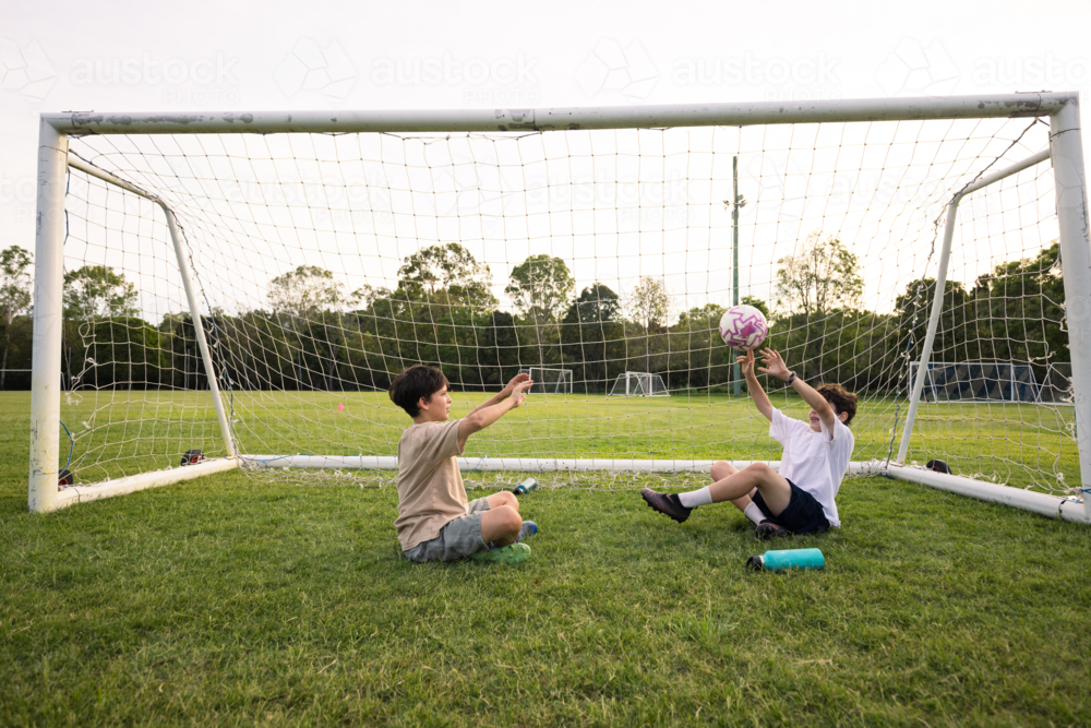 Two young boys playing with soccer ball while seated on grass field near goalpost - Australian Stock Image