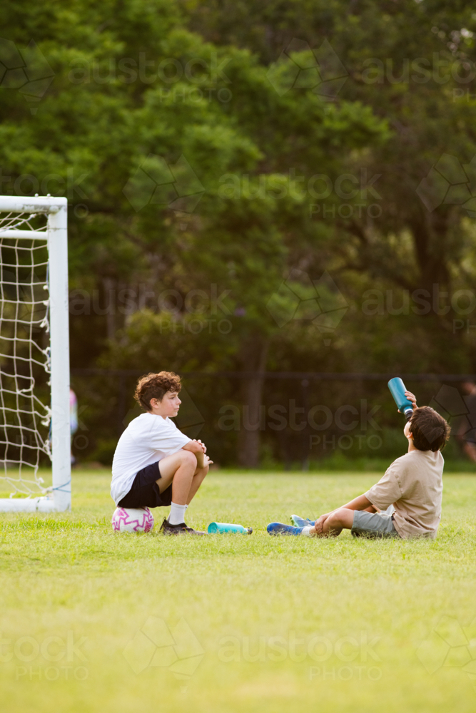 Two young boys sitting on grassy field in front of soccer goal one drinking from his drink bottle - Australian Stock Image