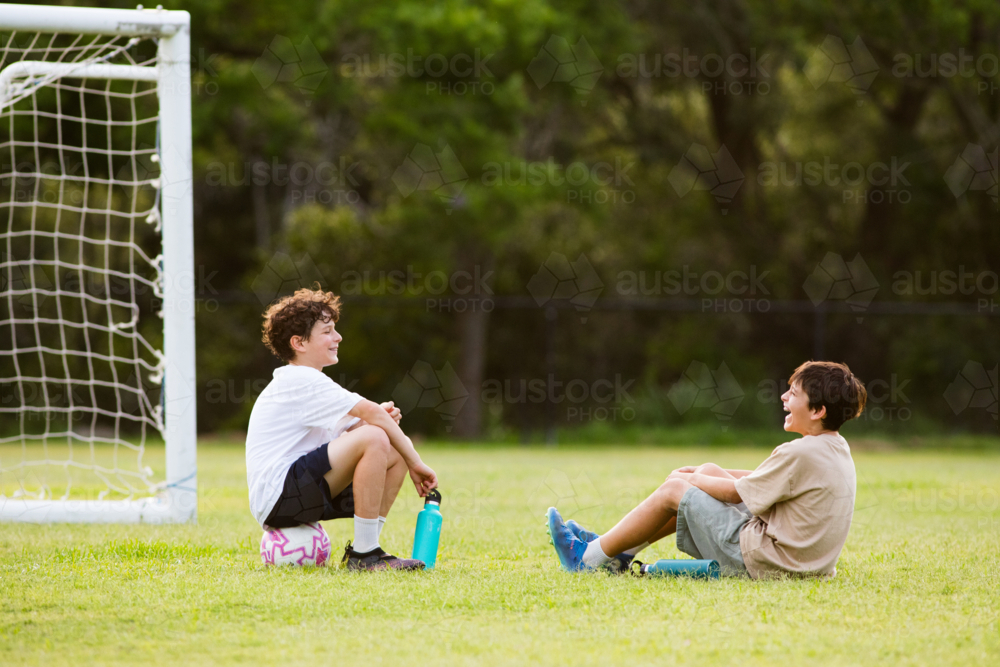 Two young boys sitting on the grassy field in front of soccer goal with their water bottles - Australian Stock Image