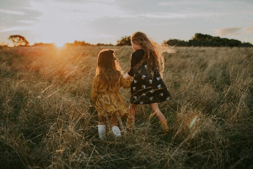 Two young fashionable sisters walking through long grass in a paddock at sunset - Australian Stock Image
