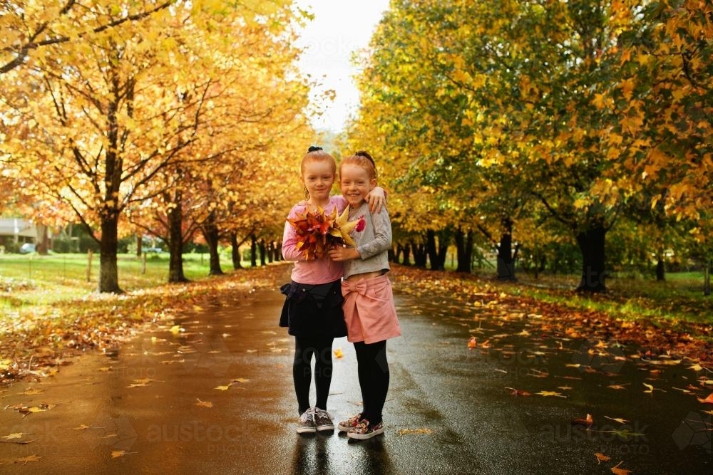 Two young girls holding autumn leaves on a tree lined street. - Australian Stock Image