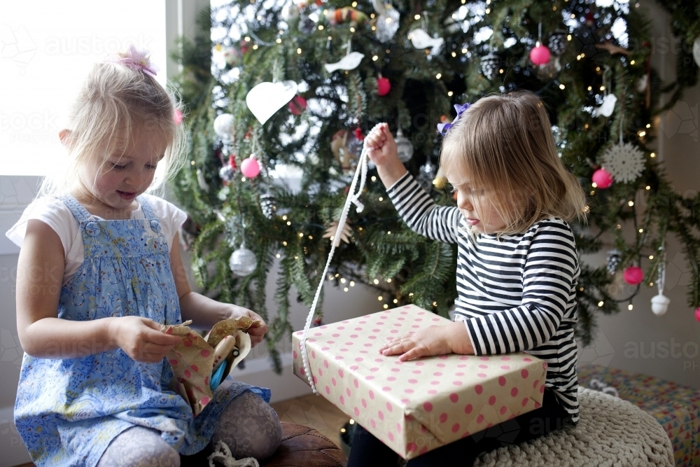Two young girls in front of Christmas tree unwrapping presents - Australian Stock Image