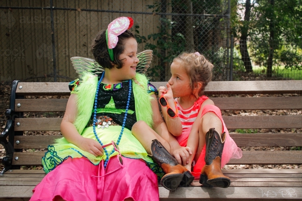 Two young girls wearing bright dress-up sitting on a park bench - Australian Stock Image