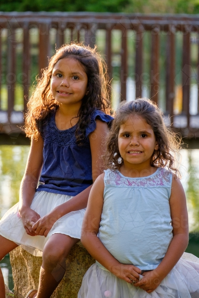 Two young girls with curly hair sitting on a huge rock outdoors looking at camera together - Australian Stock Image