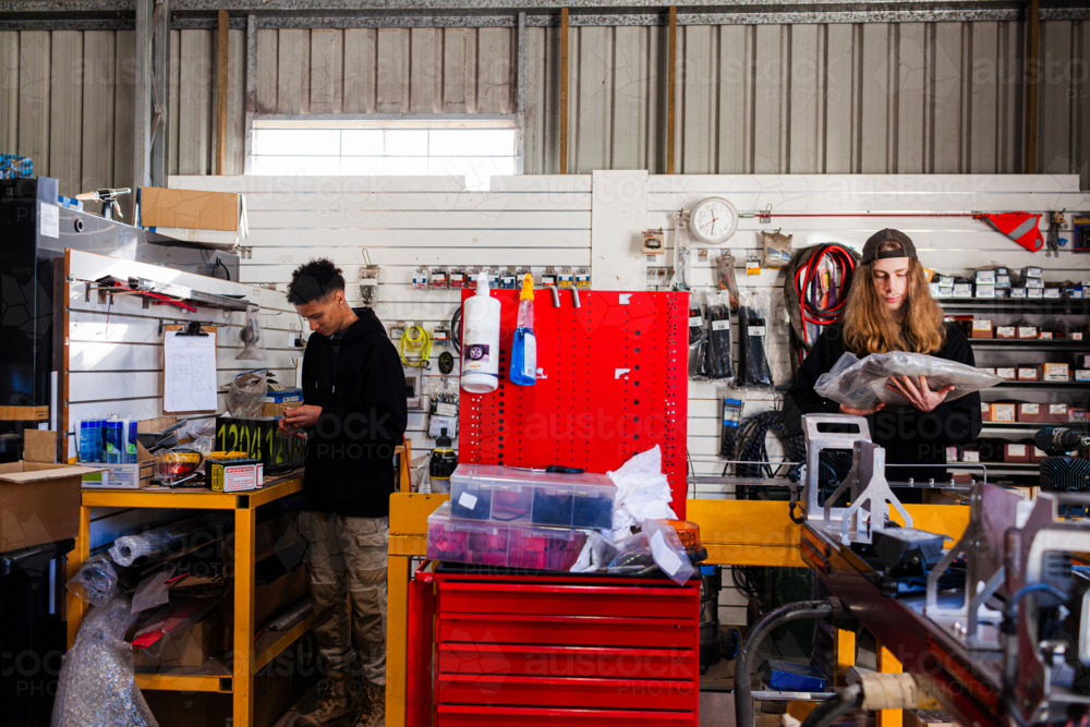 Two young male mechanics in workshop area one working on battery and one holding a new component - Australian Stock Image