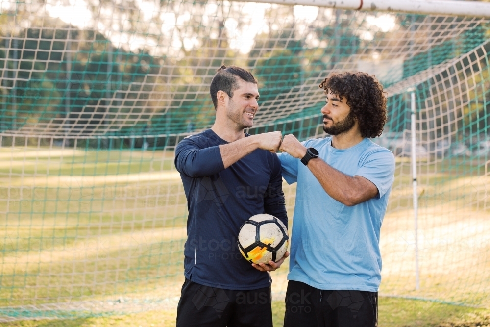 Two young men standing on the field looking at each other, bumping their fists with a soccer ball - Australian Stock Image