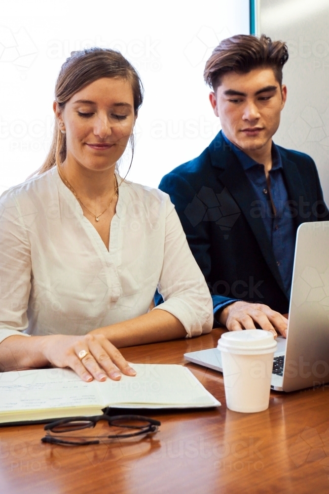 Two young professional office workers sitting at a meeting table with laptop and notebook - Australian Stock Image