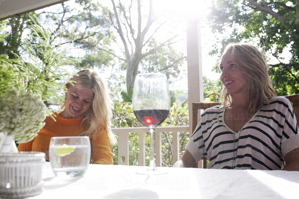 Two young women sitting at a table smiling and drinking wine in the sunshine - Australian Stock Image