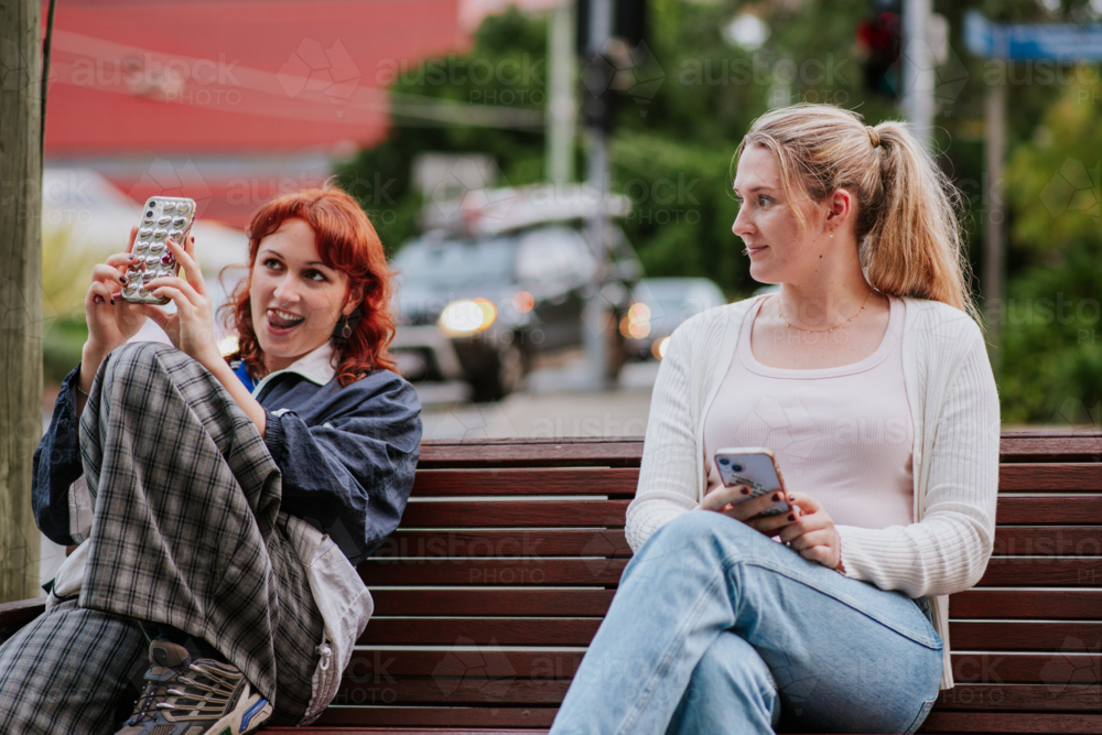 Two Young Women Sitting on a Bench Using Smartphones at a Busy Urban Intersection - Australian Stock Image
