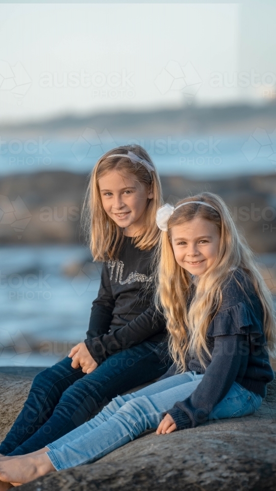 Under 10 girls sitting on rock at beach - Australian Stock Image