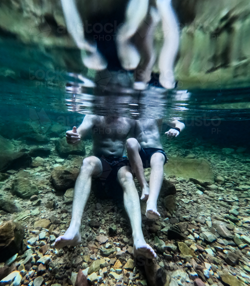 Underwater of man and boy sitting Side by side on rock with reflections with only bodies visible - Australian Stock Image