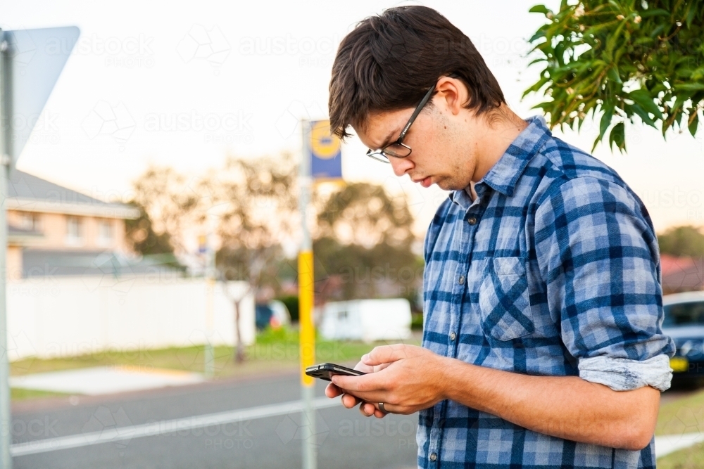 Uni student waiting at bus stop using mobile phone - Australian Stock Image