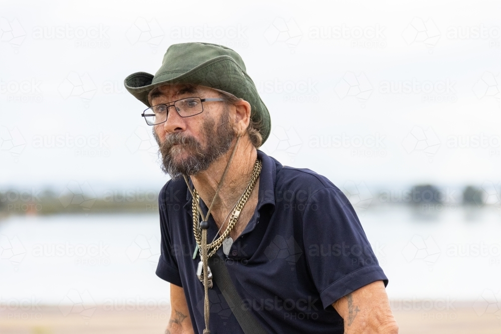 unkempt middle-aged man wears a hat and sunglasses and necklaces with light background with water - Australian Stock Image