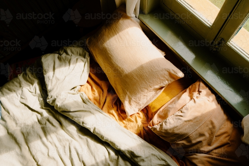 Unmade bed with crumpled sheets and sunlight coming in through the window - Australian Stock Image