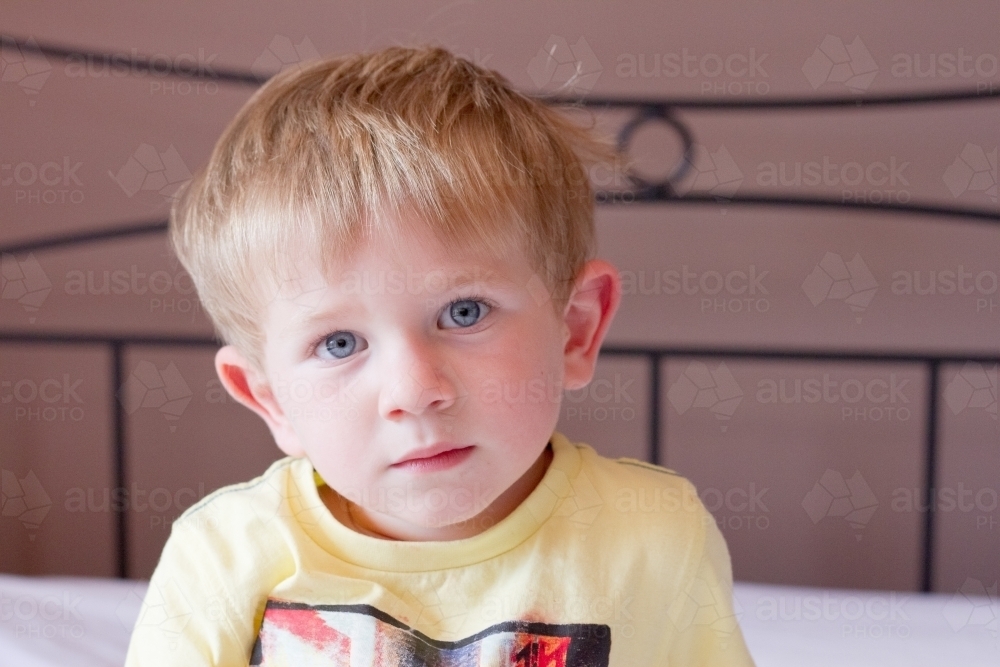 Unsmiling toddler boy sitting on bed - Australian Stock Image
