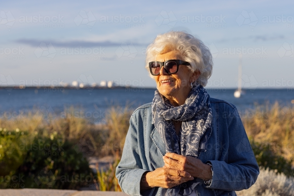 upper body of elderly lady with white hair wearing denim jacket and scarf outside near harbour - Australian Stock Image