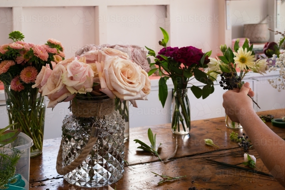 Vases of roses and other flowers on an old wooden work bench with womans hand holding arrangement - Australian Stock Image