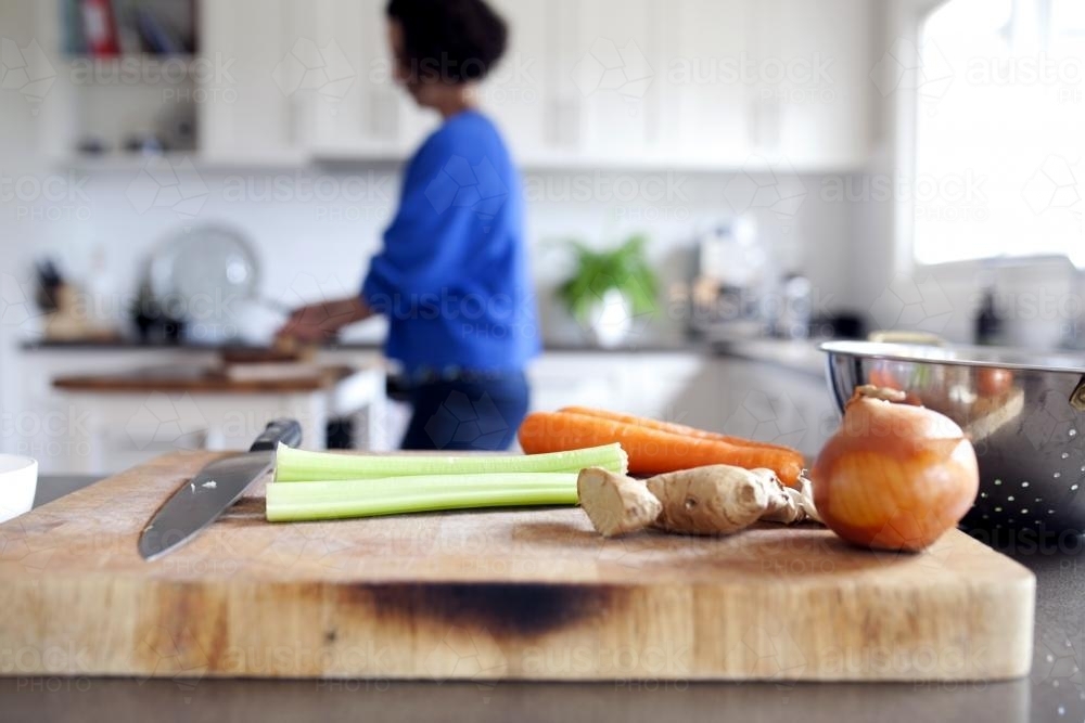 Vegetables and chopping board in kitchen with woman in background - Australian Stock Image