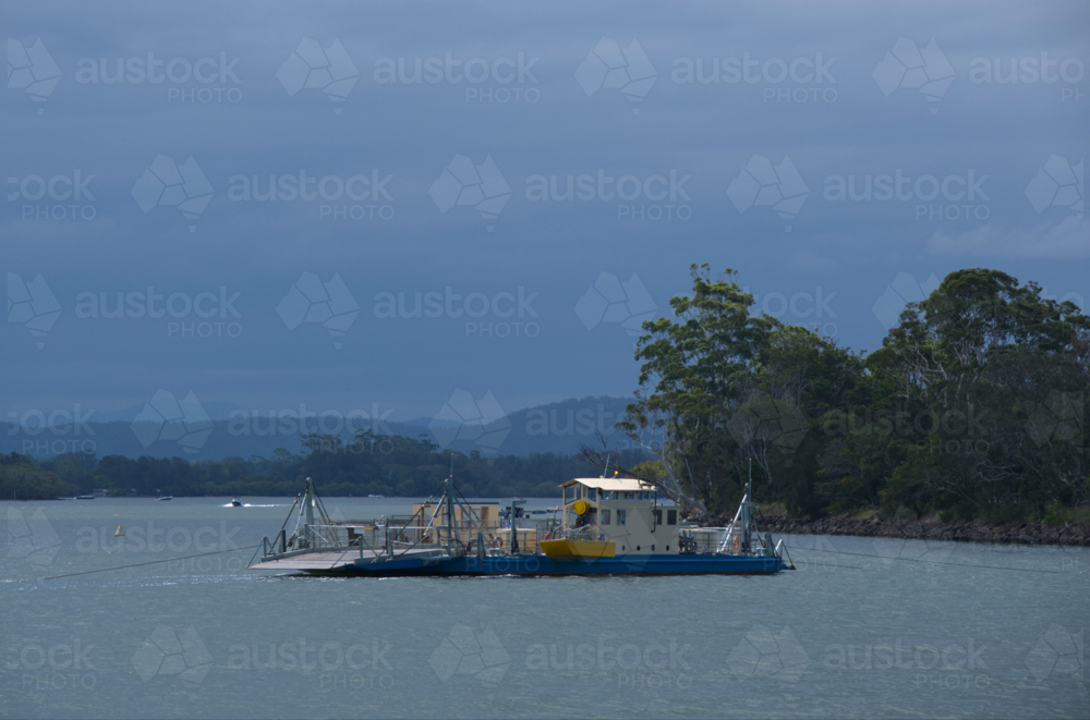 Vehicle ferry crossing river in sun, clouds and trees in background - Australian Stock Image