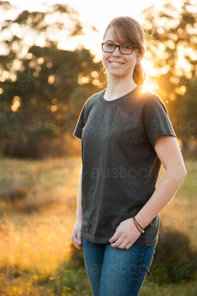 Vertical portrait of a young person with glasses smiling in rural setting with sun flare - Australian Stock Image