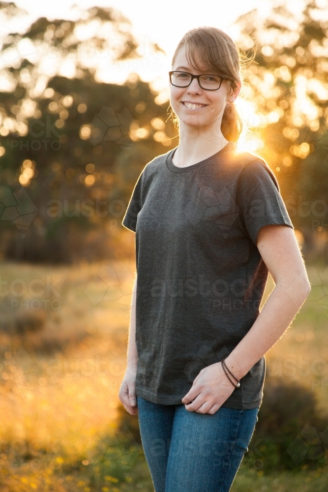 Vertical portrait of a young person with glasses smiling at camera with sun flare - Australian Stock Image
