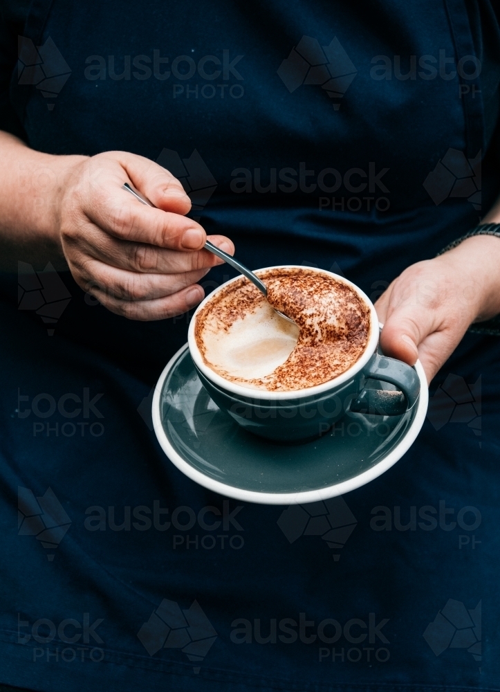 Vertical shot of a hand holding a saucer and a cup of coffee while scraping off the froth in it. - Australian Stock Image
