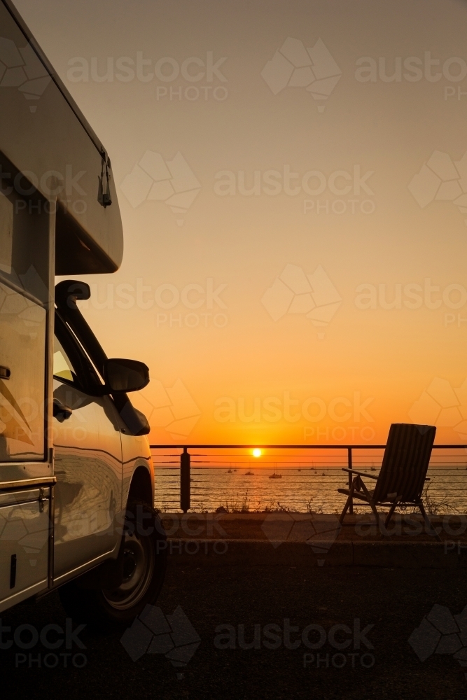 Vertical shot of a white camping van during sunset in front of the sea with foldable chair - Australian Stock Image