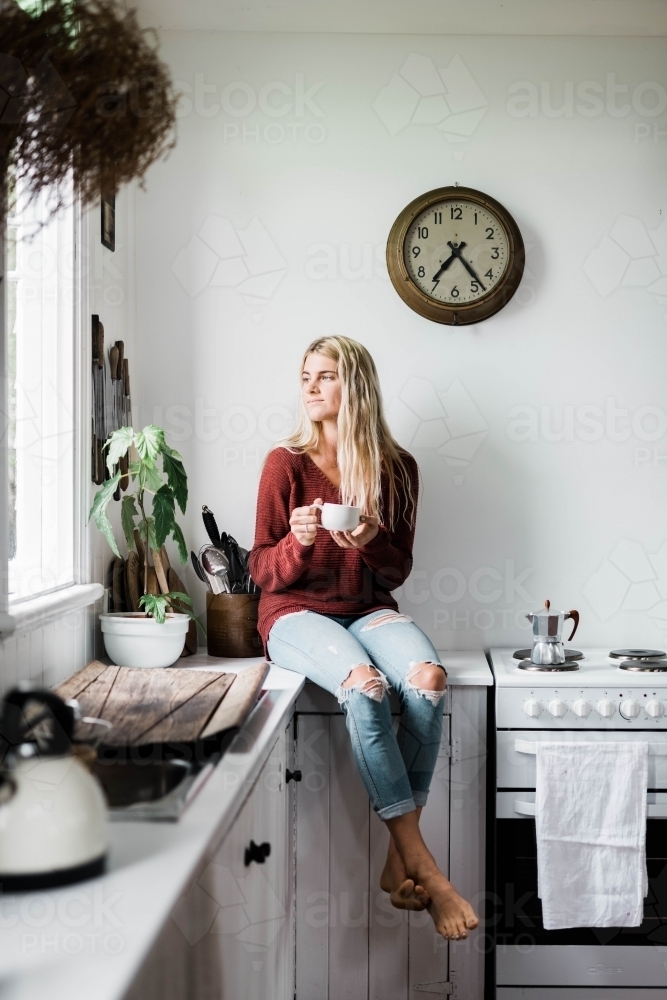 Vertical shot of a woman sitting on the kitchen counter near the window while drinking coffee - Australian Stock Image