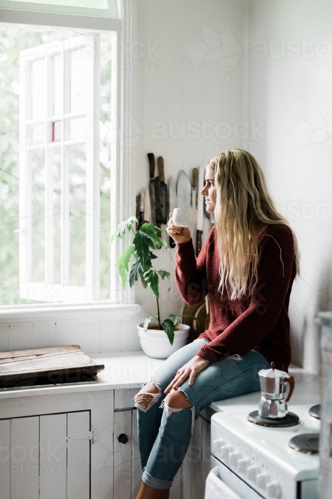 Woman sitting on the kitchen counter near the window while drinking coffee - Australian Stock Image