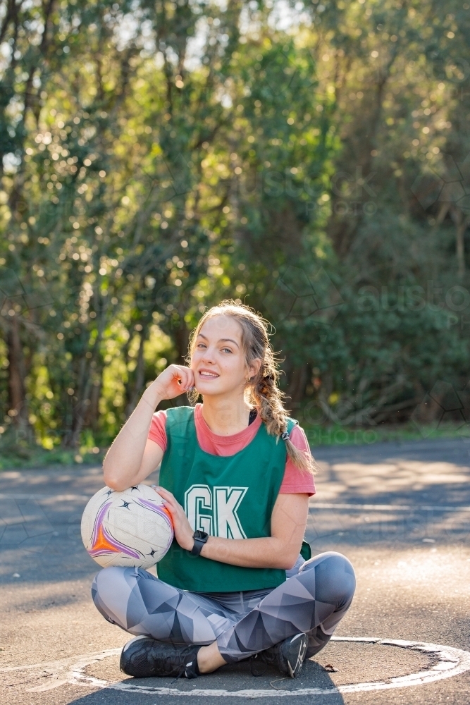 vertical shot of a young woman sitting on the ground touching her face and the net ball on her lap - Australian Stock Image