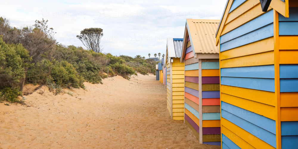 Vibrant beach huts on sandy path with greenery and cloudy sky in background - Australian Stock Image