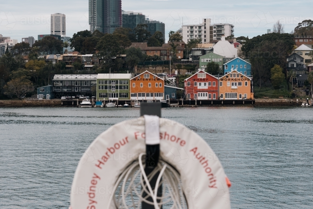 view across the harbour of colourful boat sheds on the foreshore - Australian Stock Image