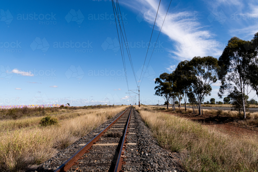 view along railway tracks lined with dry grass and trees and cotton bales in distance - Australian Stock Image
