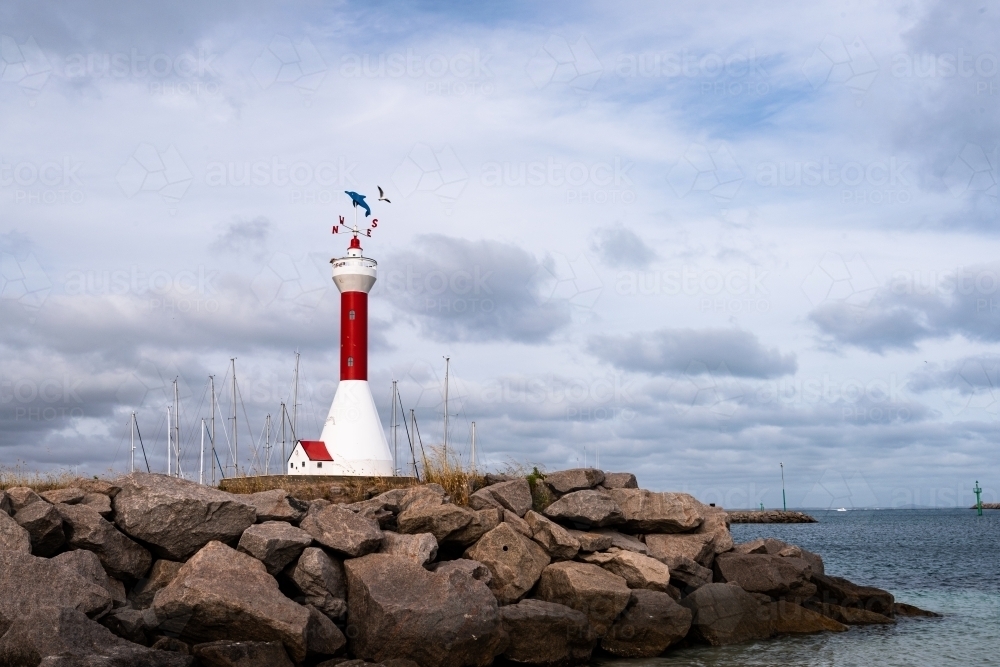 View of a miniature lighthouse on a rocky groyne with dramatic sky and harbour - Australian Stock Image
