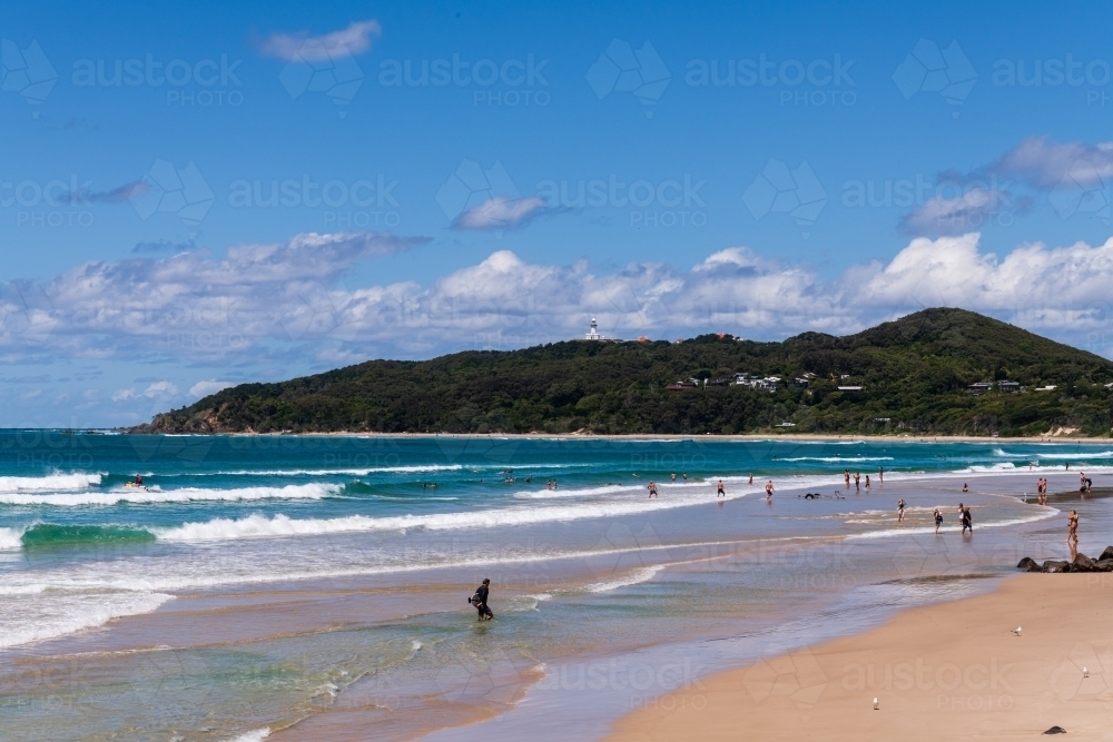 View of Cape Byron and lighthouse from the beach at Byron Bay with surfers, sandy foreshore - Australian Stock Image