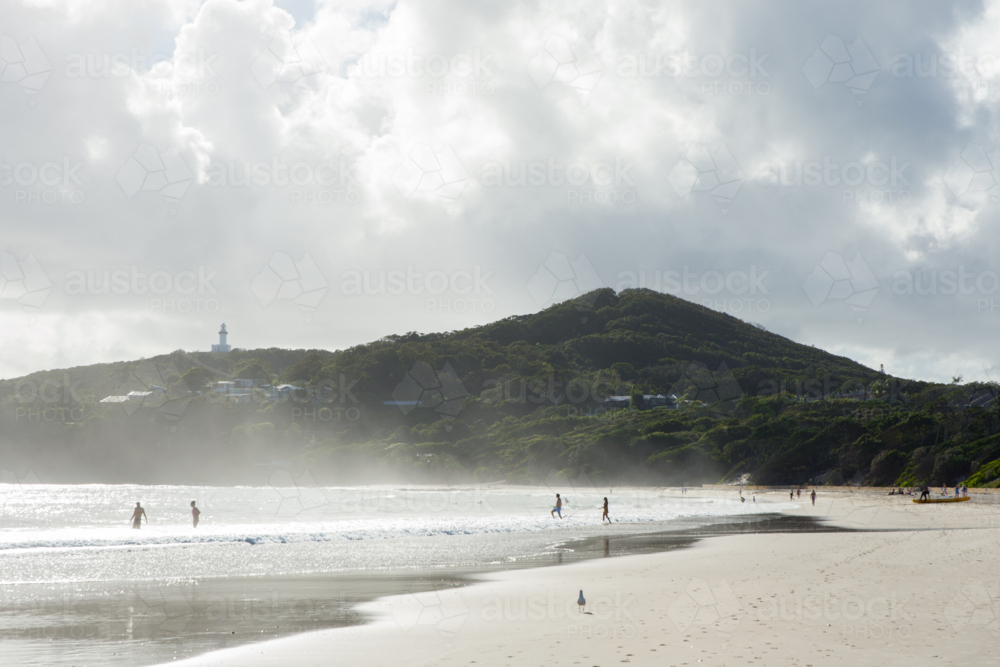 View of Cape Byron and lighthouse from the beach with swimmers and sandy foreshore - Australian Stock Image