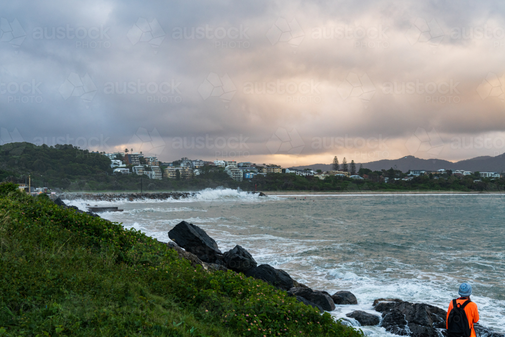 View of harbour with waves crashing on breakwater and person in foreground with backpack - Australian Stock Image