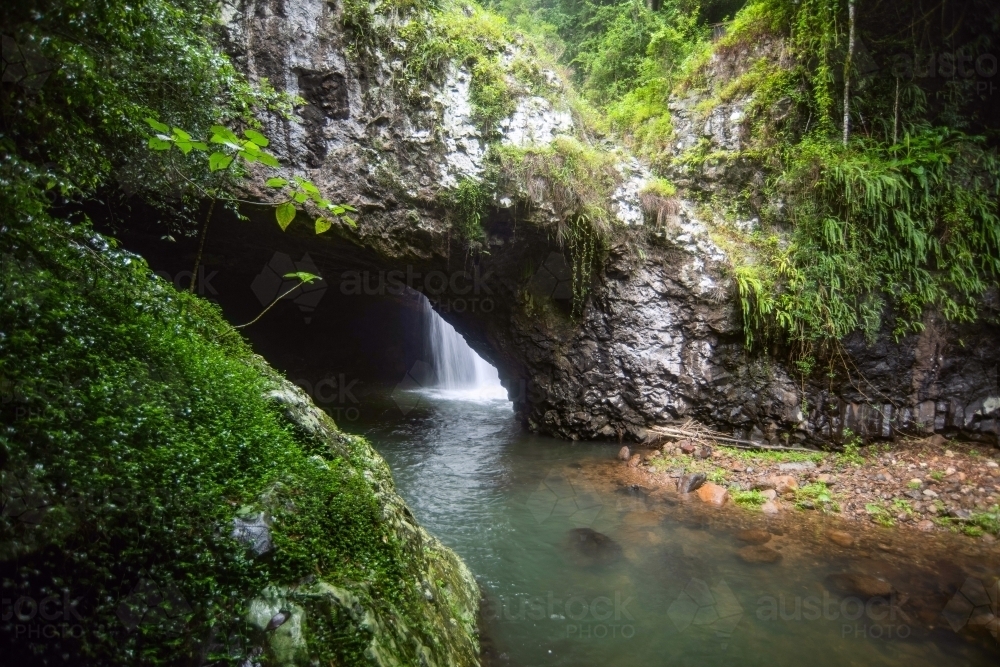 View of the waterfall through an opening of a cave mouth with the greenery surrounds - Australian Stock Image