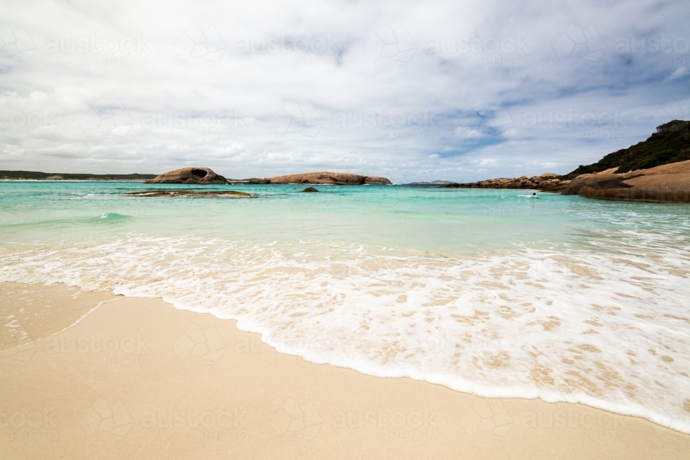 View of water patterns on beach of a pretty cove with swimmer in the distance and islands - Australian Stock Image