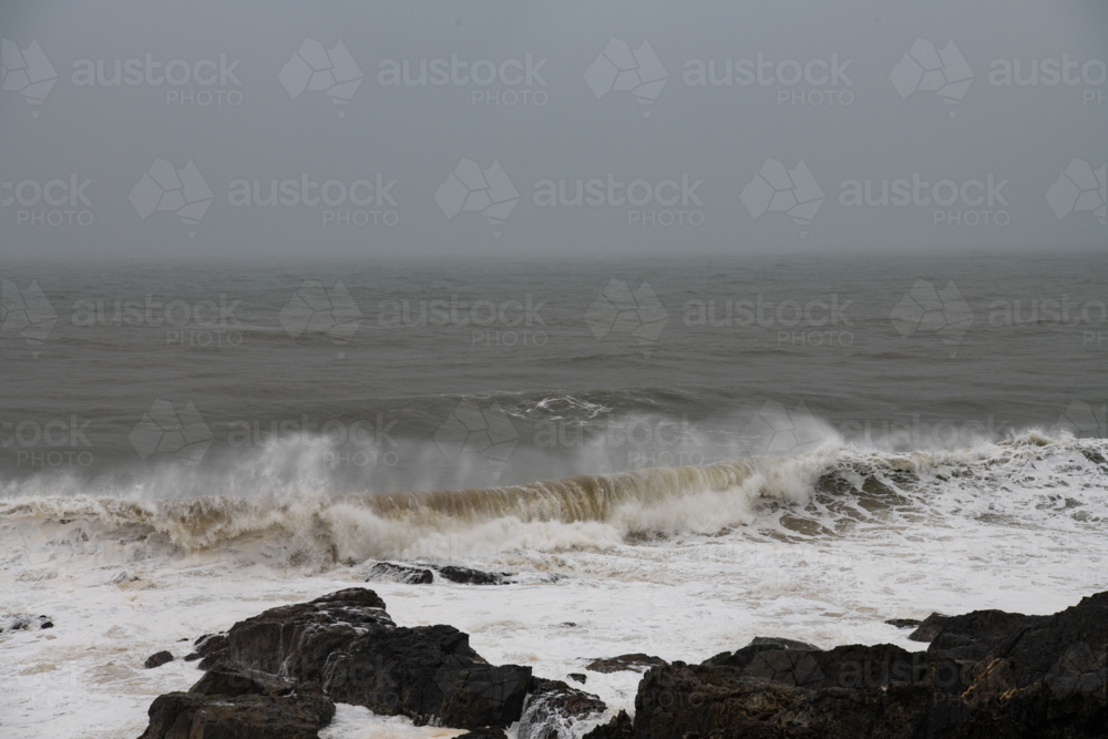 View of wave breaking with spray and froth,  and rocks in the foreground - Australian Stock Image