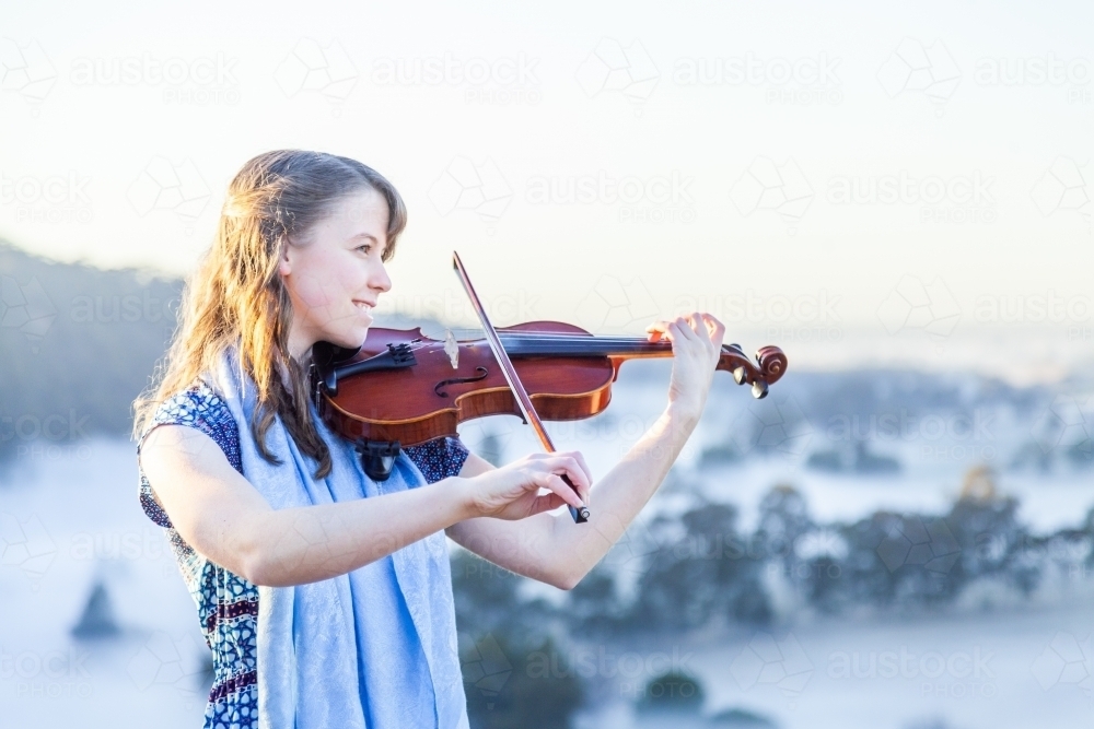 Violin player standing out on cliff top playing music on misty morning - Australian Stock Image