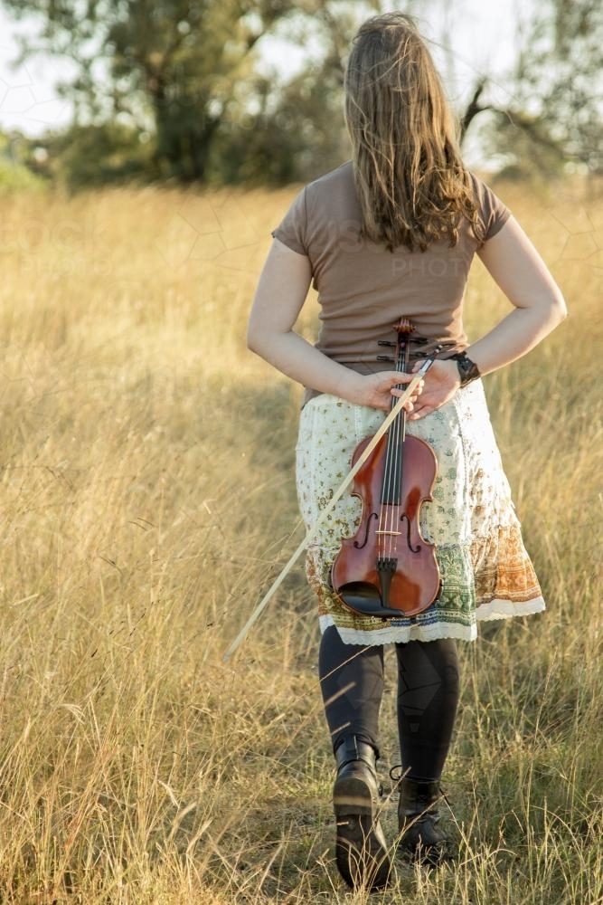 Violinist holding violin behind her and she walks through a paddock - Australian Stock Image