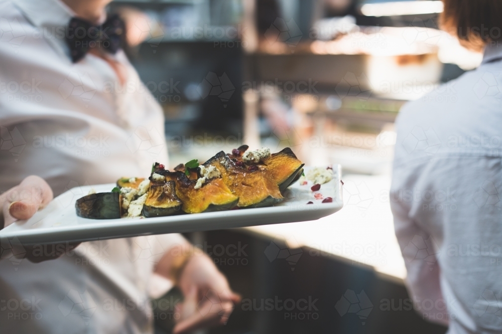 Waiter wearing a bow tie and carrying a plate of roasted pumpkin in a restaurant kitchen - Australian Stock Image