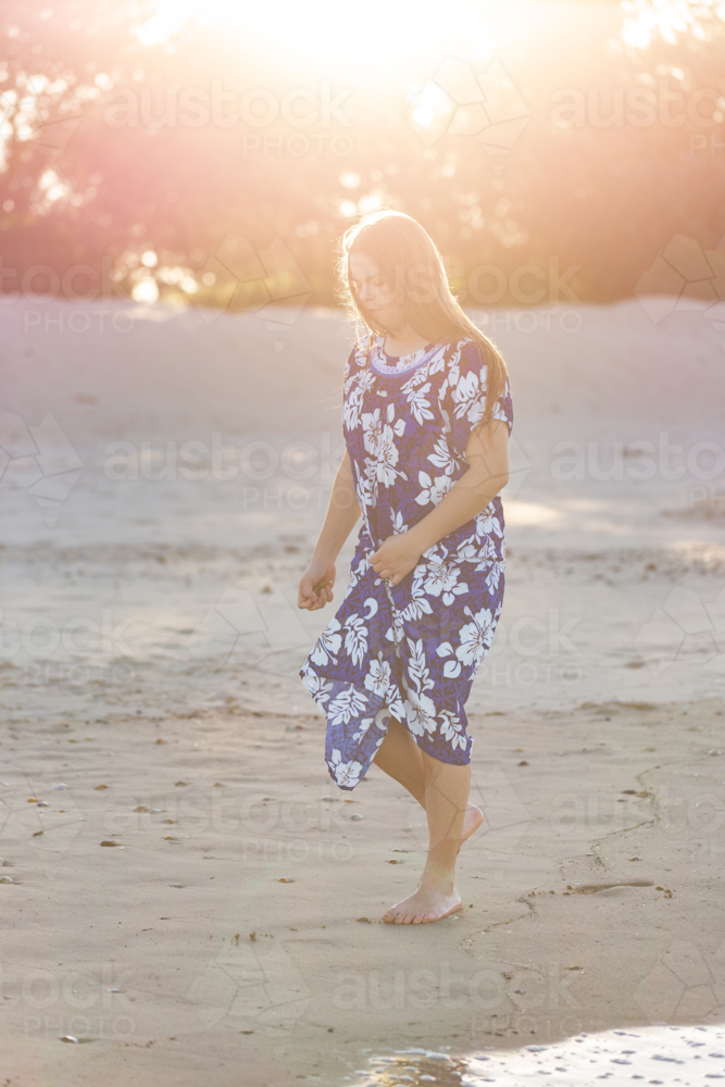 Warm sun haze glow over teen Torres Strait Islander girl walking along beach in floral dress - Australian Stock Image