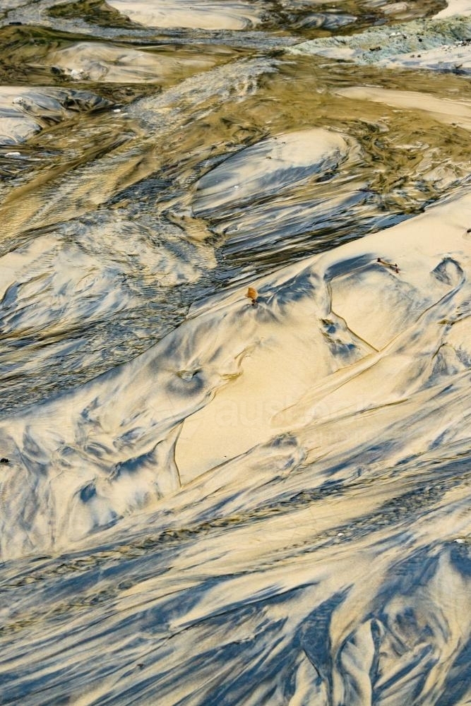 Water running over patterns in wet yellow and black mineral sands on a beach at low tide - Australian Stock Image