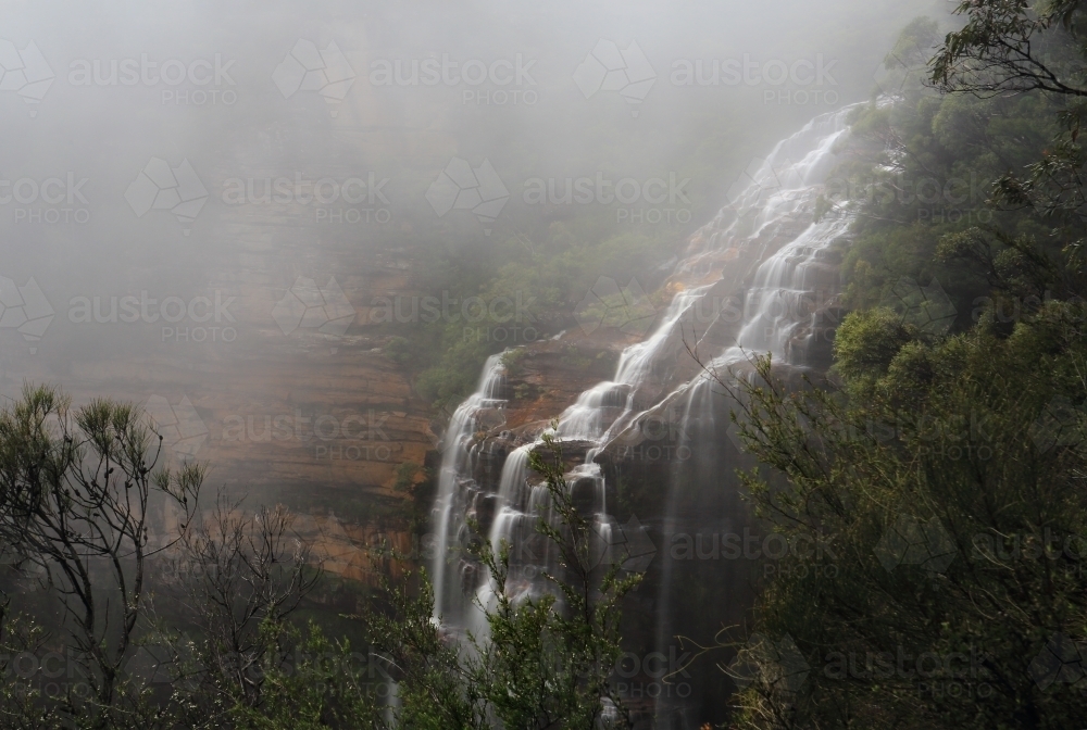 Waterfall flowing down the cliff face  with a heavy dense fog subduing visibility - Australian Stock Image