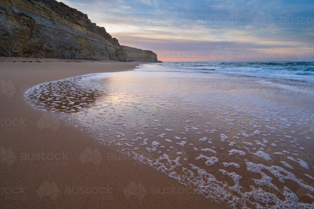 Wave patterns left on wet sand below rugged sea cliffs at twilight - Australian Stock Image